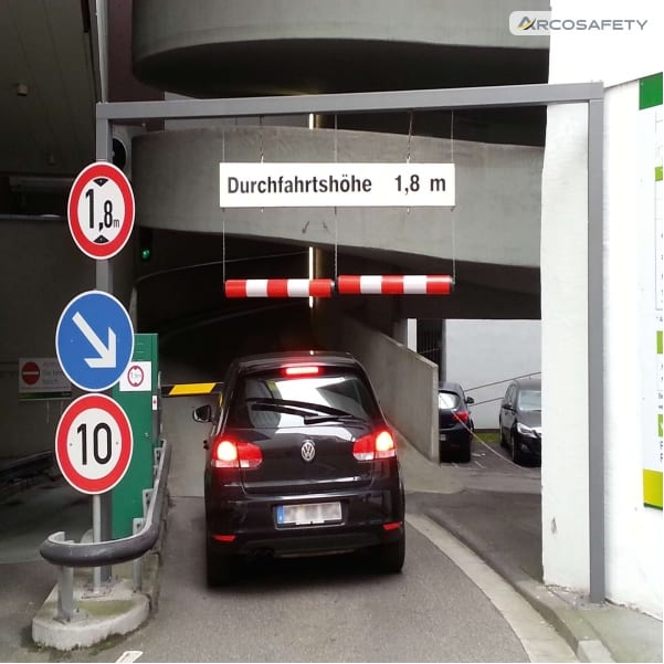 Ingresso di un parcheggio multipiano con due limitatori pendenti in plastica MORION rossi e bianchi sospesi a un portale grigio, cartello di altezza massima 1,8 m e un'auto in entrata