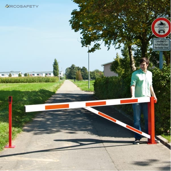 Sbarra girevole COMPACT con asta bianca a bande rosse e traversino diagonale di rinforzo, manovrata da un uomo su una stradina asfaltata, con montante di appoggio rosso e cartello di divieto di transito ai veicoli a motore
